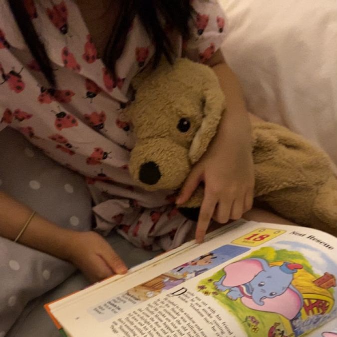 Uma pessoa está sentada na cama, segurando um livro e um brinquedo de pelúcia, enquanto lê.