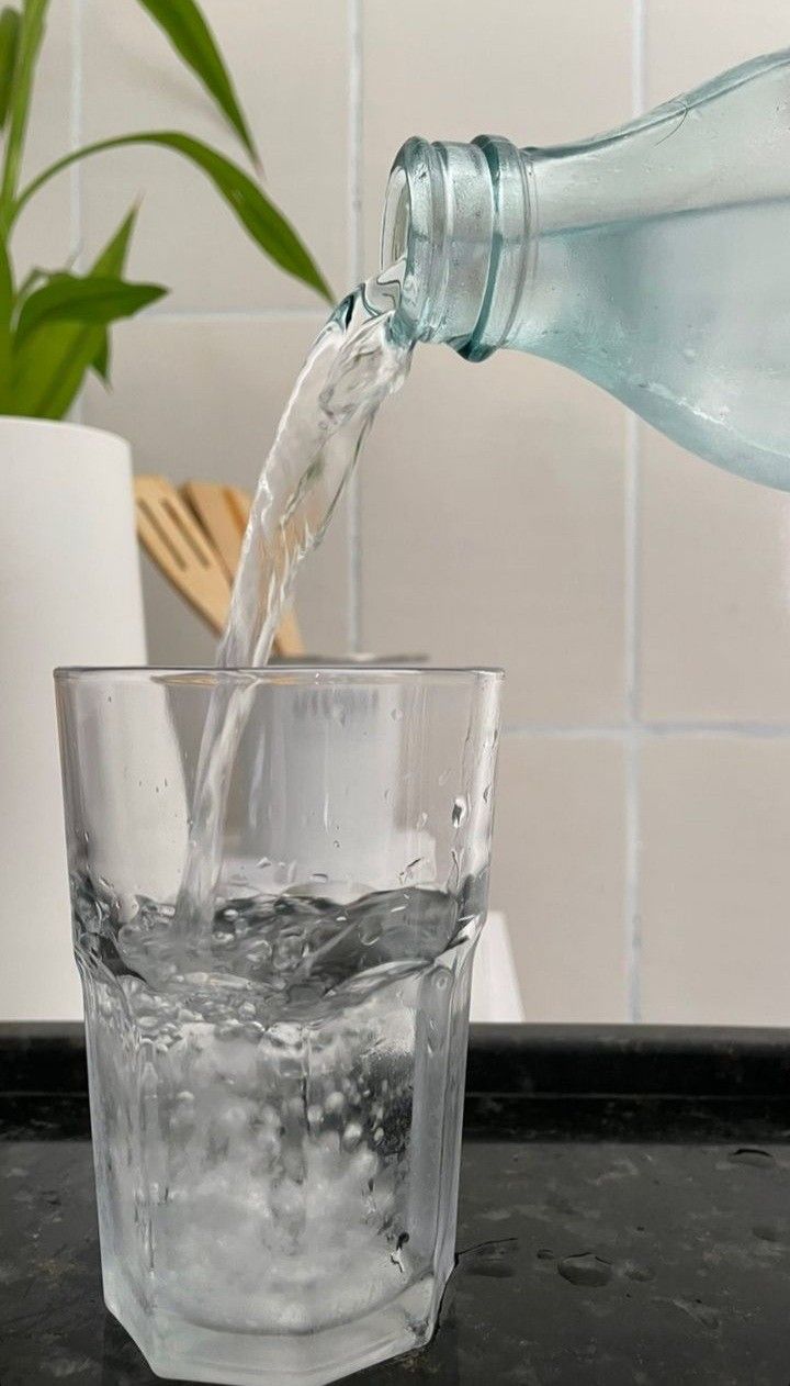Uma pessoa está despejando água de uma garrafa em um copo transparente sobre uma superfície escura.