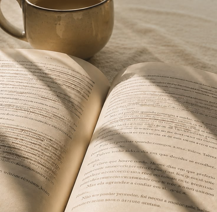 Um livro aberto com páginas escritas, ao lado de uma xícara sobre uma superfície clara, com sombras visíveis.